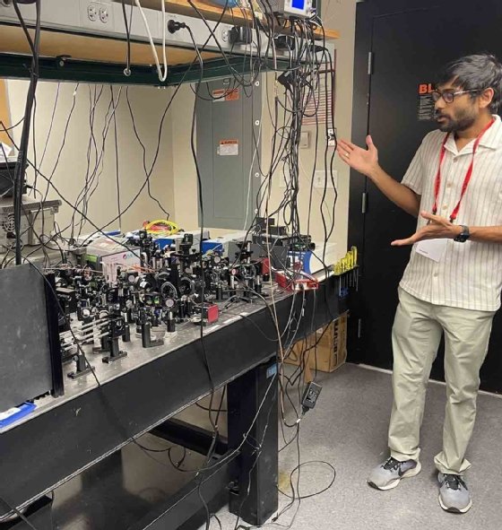 Atanu Ghosh stands to the right and gestures toward a table of plugs, wires and quantum technologies while leading a tour of MIT's quantum labs. 