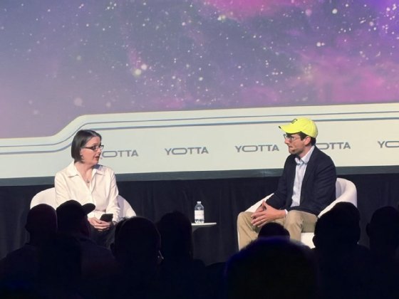 The image shows two people, the one on the left in a white button-down shirt and on the left in a dark sport coat, light colored button-down shirt and yellow baseball cap, seated on a stage in front of a backdrop that says 