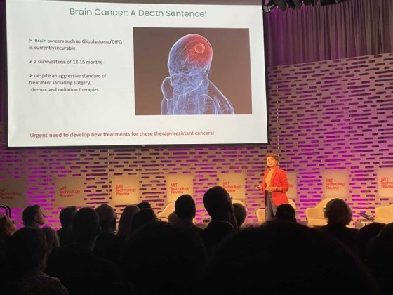 A crowd of seated people watches Deblina Sakar, who stands on stage in front of a PPT slide labeled 'Brain Cancer: A Death Sentence!' at MIT EmTech Future. 