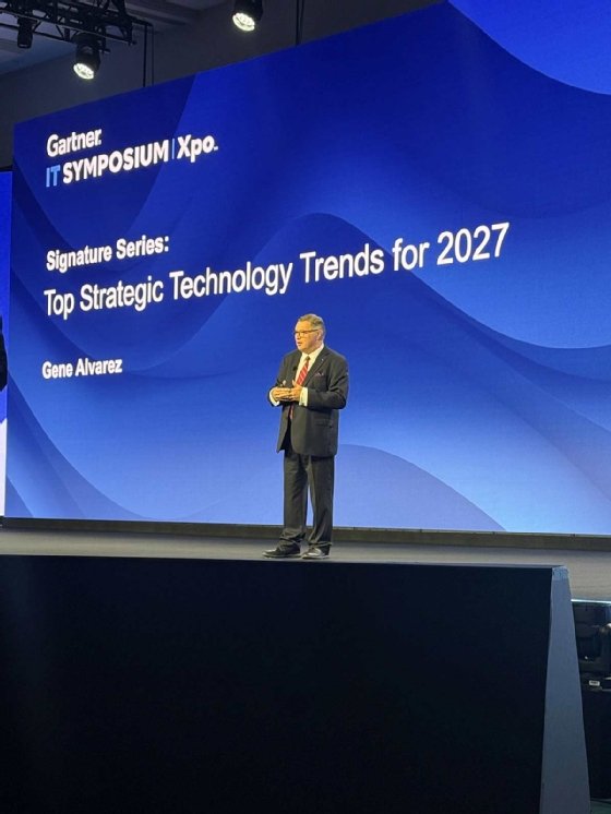 An image of Gene Alvarez in a suit speaking onstage at the Gartner IT Symposium, with a blue background.