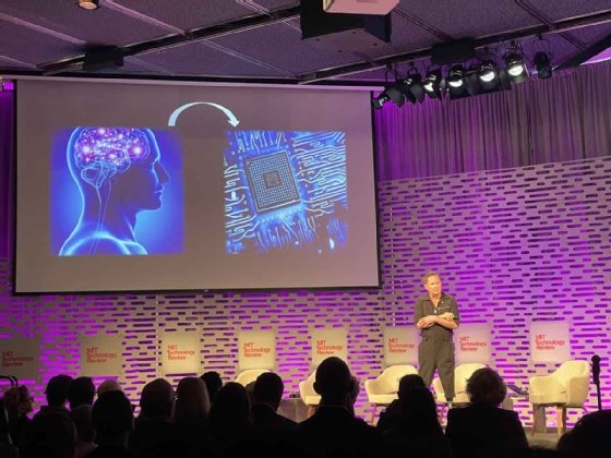 A crowd of seated people watches Hugh Herr, who stands on stage in front of a PPT slide showing a human brain with an arrow pointing to a technology grid at MIT EmTech Future. 