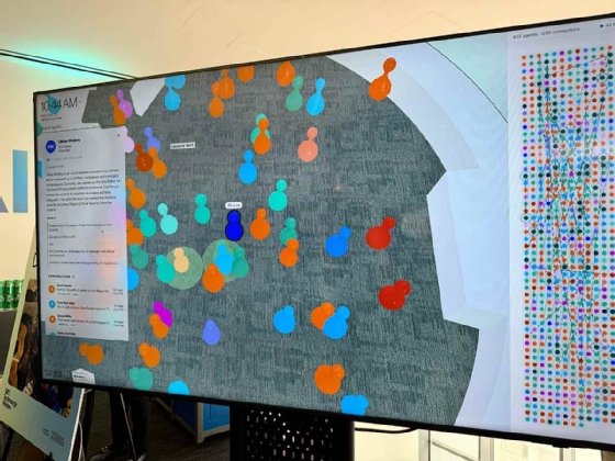 TV monitor showing colorful objects on a simulated conference floor, each representing an attendee's agent twin. The left-hand side shows the bio for 