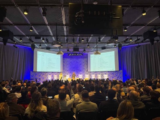 Image showing MIT Media Lab stage with Gorskikh (left) and Bhattacharya (right) standing in front of a PPT slide showing agent-building steps. Many attendees sitting in front of the stage have their hands raised.