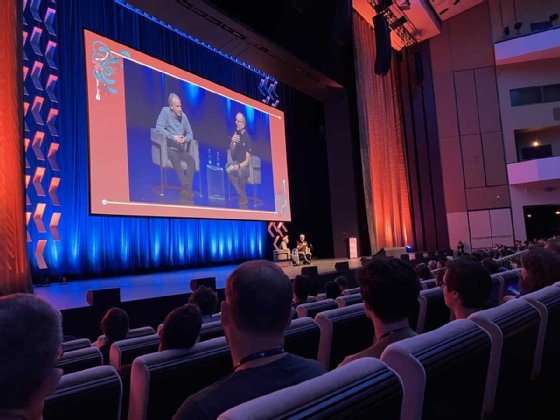 The image shows two people seated on a stage in front of an auditorium with an audience present, with their image projected larger on a screen behind them. 
