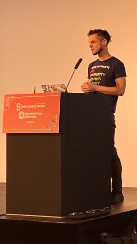 A person standing behind a conference podium in a black t-shirt with writing in white, red, orange, yellow and green. Legible words on the shirt include 