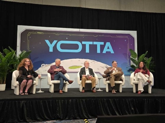The image shows five people seated on a black stage in white chairs with a backdrop that says 'YOTTA' in big letters against a graphic of the moon and bluish-purple space.